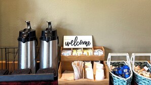A framed artwork of a basket with fruits, a welcome sign, coffee makers, and baskets of snacks. - Fidalgo Country Inn (Anacortes)