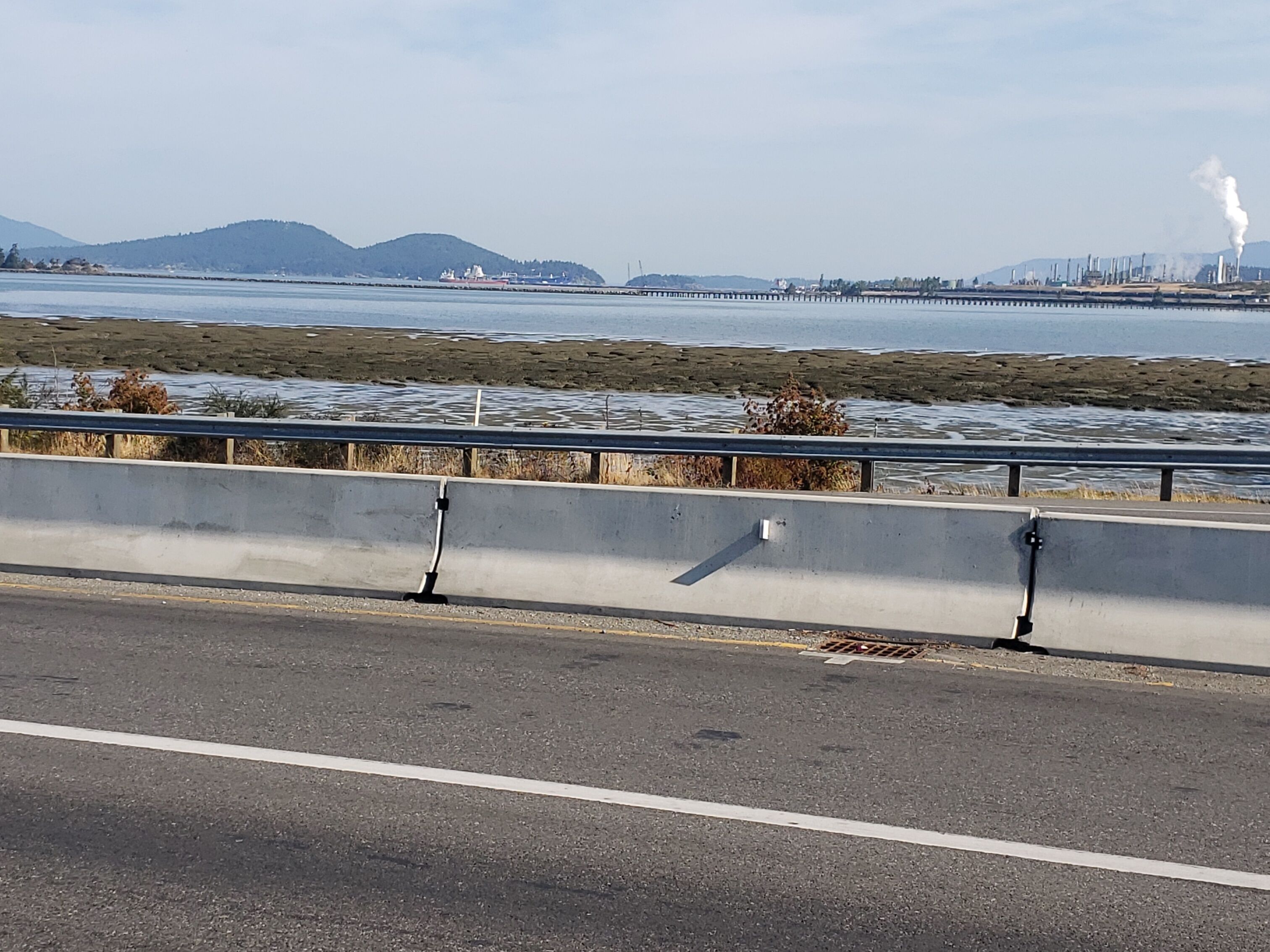 A coastal road with a concrete barrier, a body of water, and industrial structures in the distance.