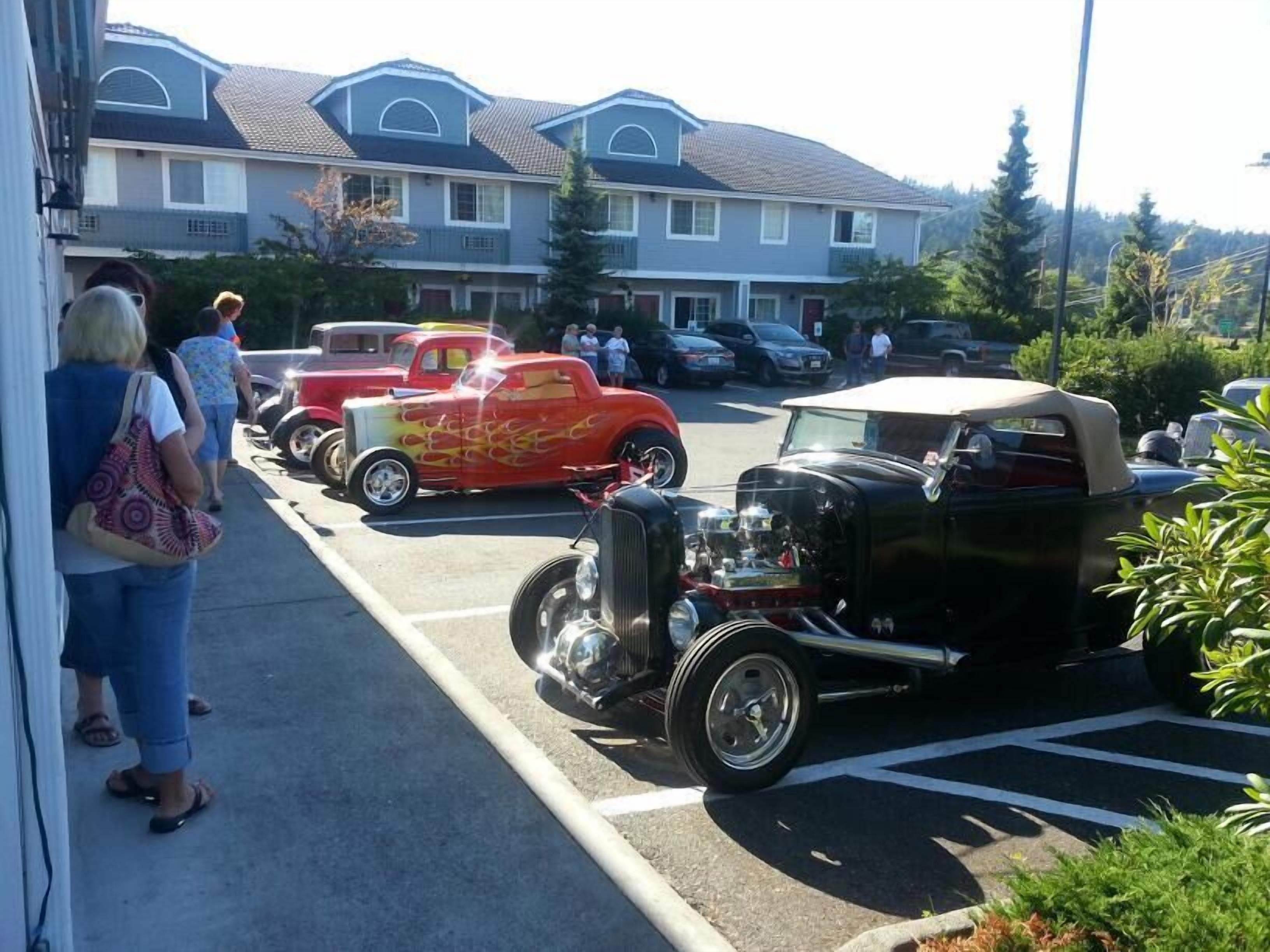 Ein Parkplatz mit Oldtimern, ein Gebäude mit Satteldach und Personen, die daneben stehen.