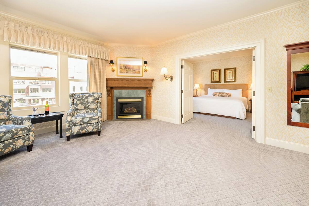 A hotel room with a fireplace, a bed, two armchairs, and a view of the city through large windows.