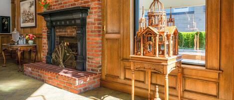 A room with a brick fireplace, a wooden table with a model, and a large window with a view of greenery.