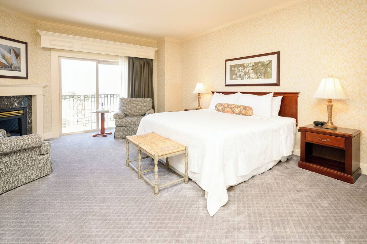 A hotel room with a large bed, two bedside tables with lamps, a chair, a small table, and a fireplace.