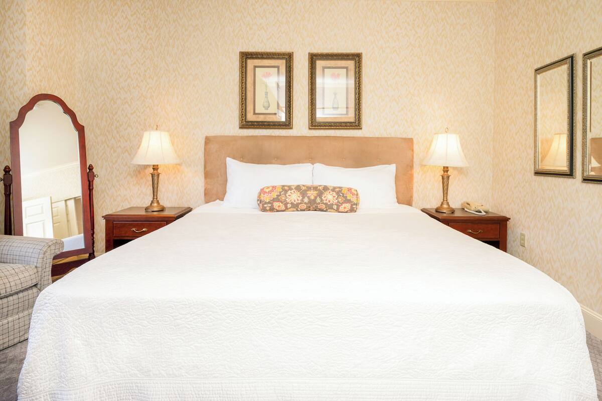 A neatly made bed with a white quilt, two bedside lamps, a floral pillow, and a mirror on the left.