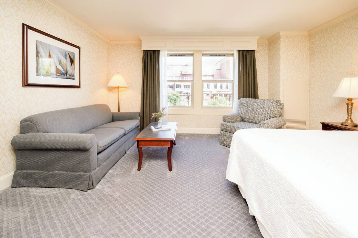 A hotel room with a grey sofa, a small wooden table, a grey armchair, a white bed, and a window with a view of buildings.