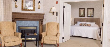 A hotel room with a fireplace, two armchairs, a bed with a white bedspread, and two framed pictures on the wall.