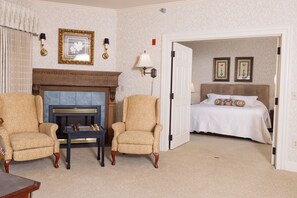 A hotel room with a fireplace, two armchairs, a bed with a white bedspread, and two framed pictures on the wall.