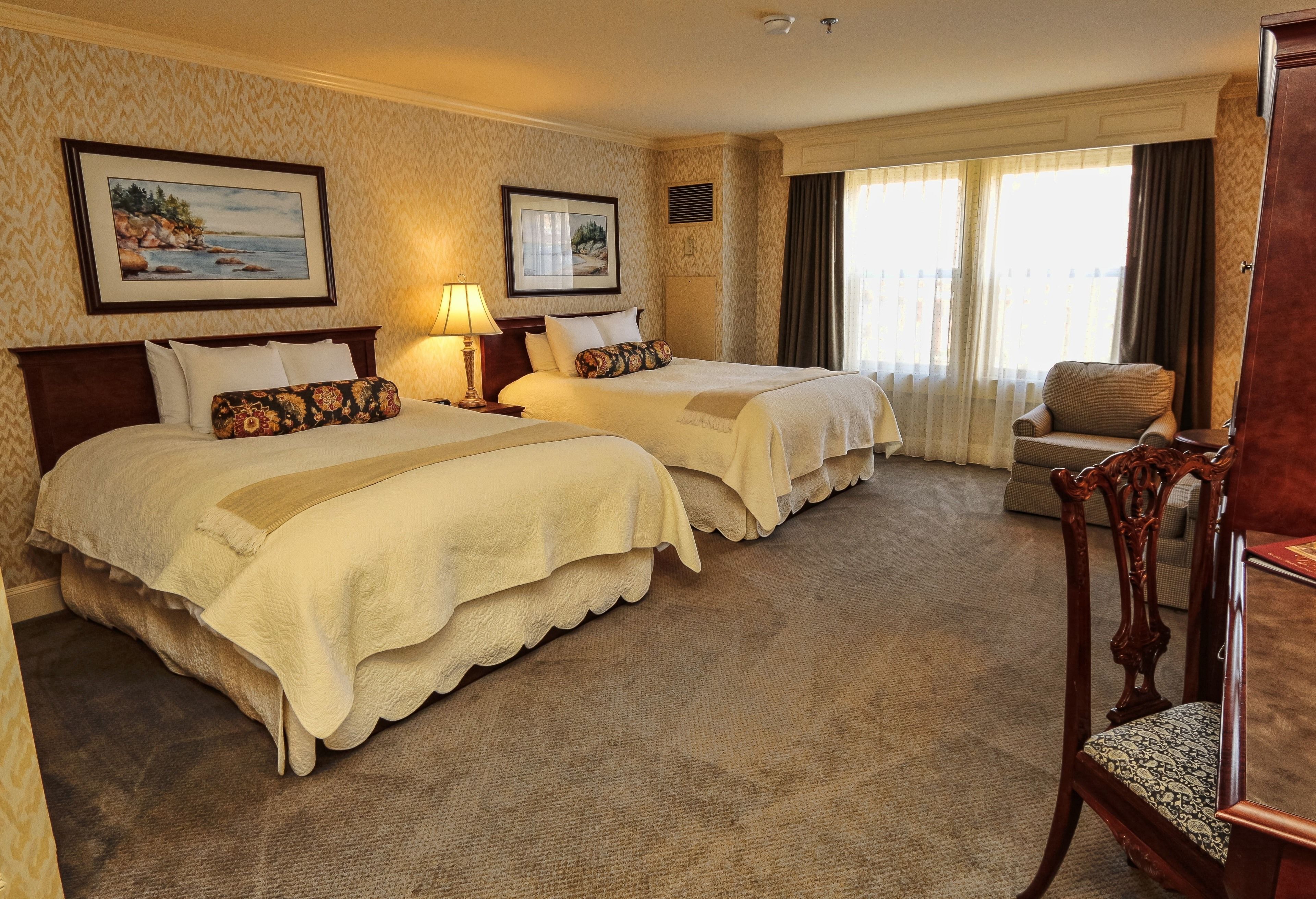 A hotel room with two beds, a wooden chair, a desk, and a large window with curtains.