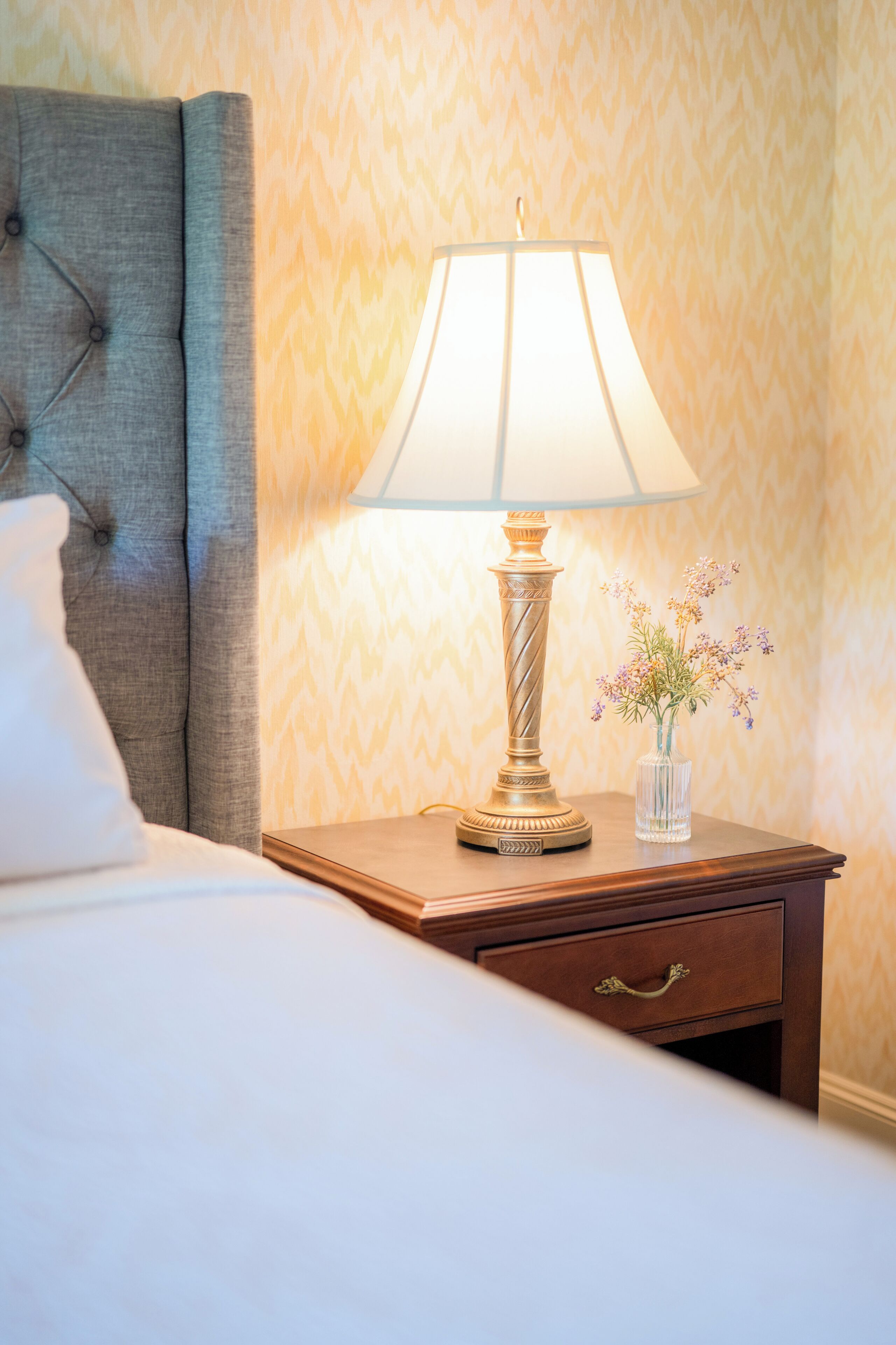 A neatly made bed with a grey headboard, a bedside lamp, and a small vase with flowers on a wooden nightstand.