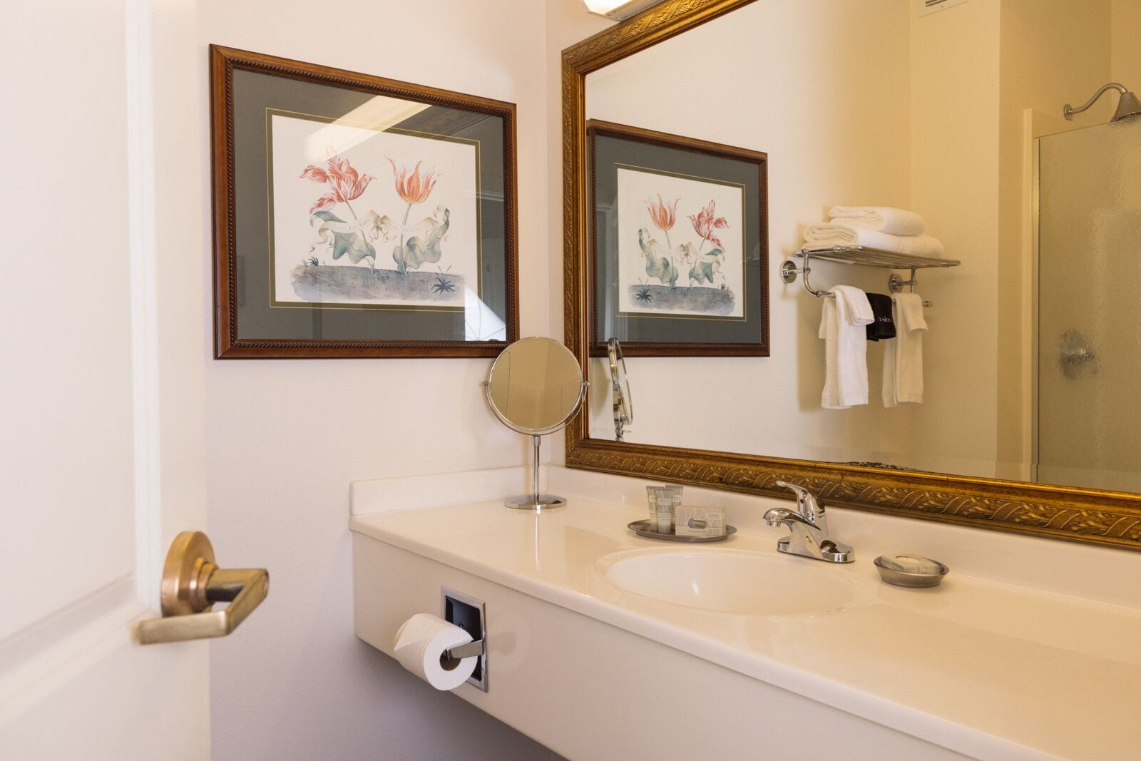 A bathroom with a large mirror, framed floral artwork, a roll of toilet paper, and a towel rack with folded towels.