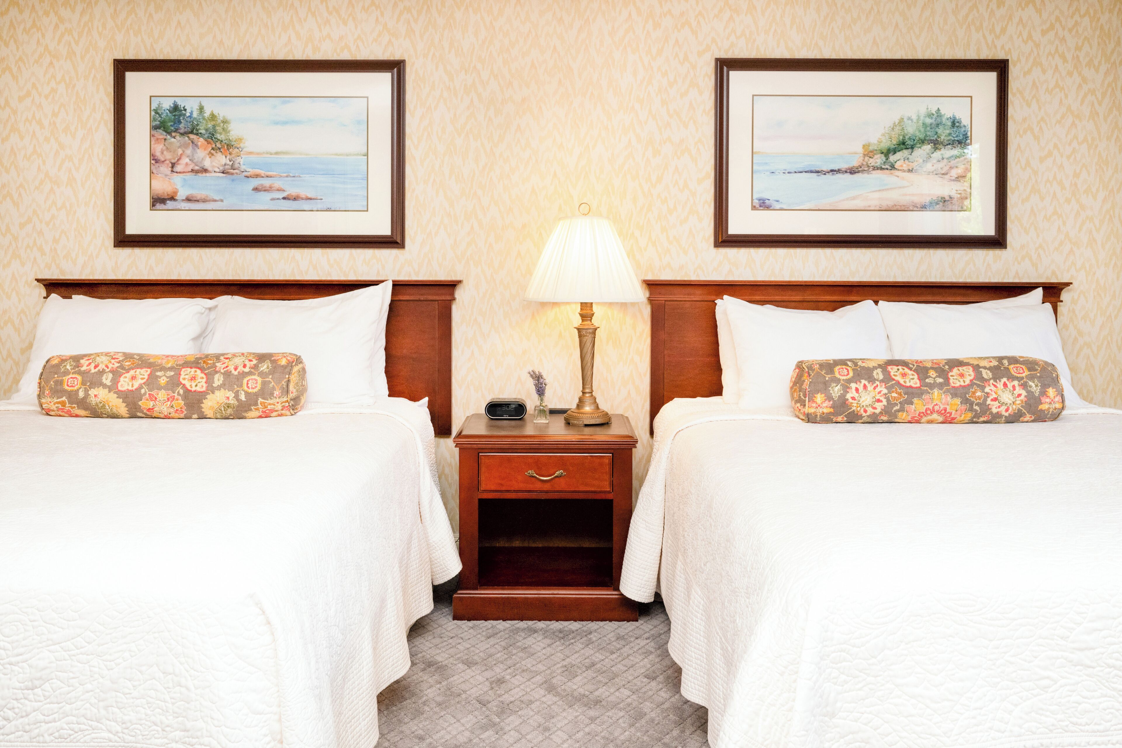 A hotel room with two beds, each with floral-patterned pillows, a nightstand with a lamp, and framed coastal landscape paintings on the wall.