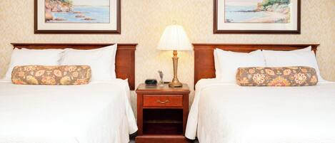 A hotel room with two beds, each with floral-patterned pillows, a nightstand with a lamp, and framed coastal landscape paintings on the wall.