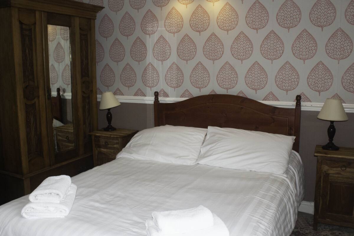 A neatly made bed with white linens and towels, a wooden headboard, and a wall with a leaf pattern.