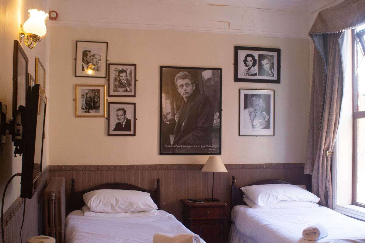 A hotel room with two single beds, a bedside table with a lamp, a radiator, and framed pictures on the wall.