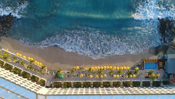 Vista aérea de un complejo turístico en la playa con filas de tumbonas y sombrillas, una piscina y un edificio de fachada azul.