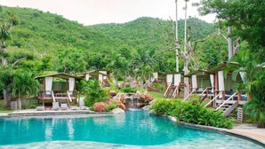 Un área de piscina con una cascada, rodeada de sillones de madera y zonas de estar cubiertas.