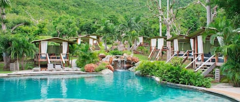 Un área de piscina con una cascada, rodeada de sillones de madera y zonas de estar cubiertas.
