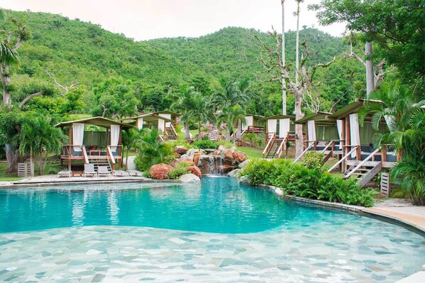 A pool area with a waterfall feature, surrounded by wooden deck chairs and covered seating areas.