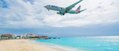 Un avión con la marca 'American Airlines' volando sobre una playa de aguas turquesas cristalinas y una orilla arenosa.