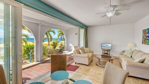 Amplia sala de estar con un ventanal que da a un jardín tropical y al océano. Hay dos sofás, una mesa de centro y un televisor sobre un mueble.