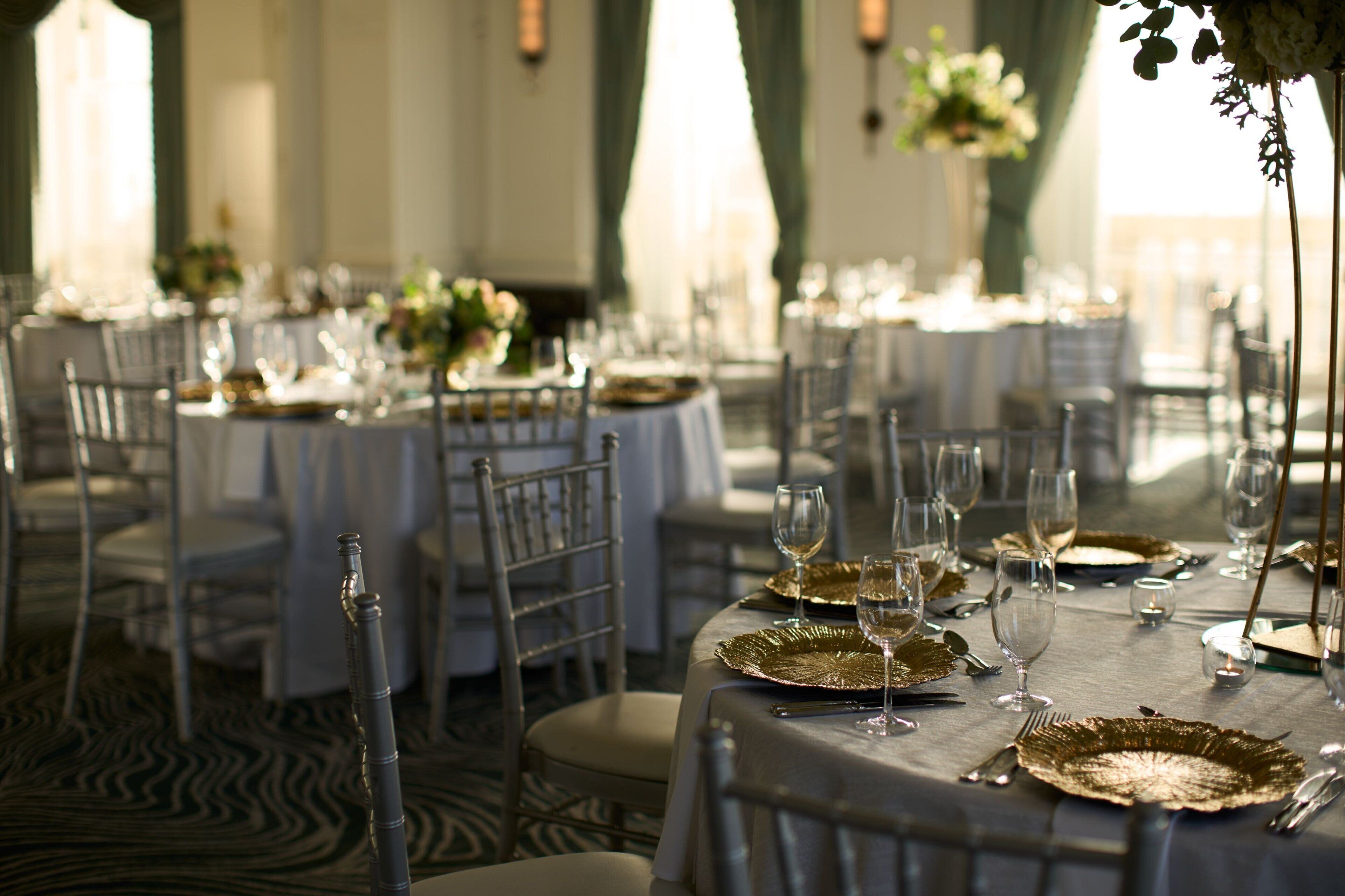 A banquet hall with round tables set for a formal event, featuring gold-rimmed plates and wine glasses.