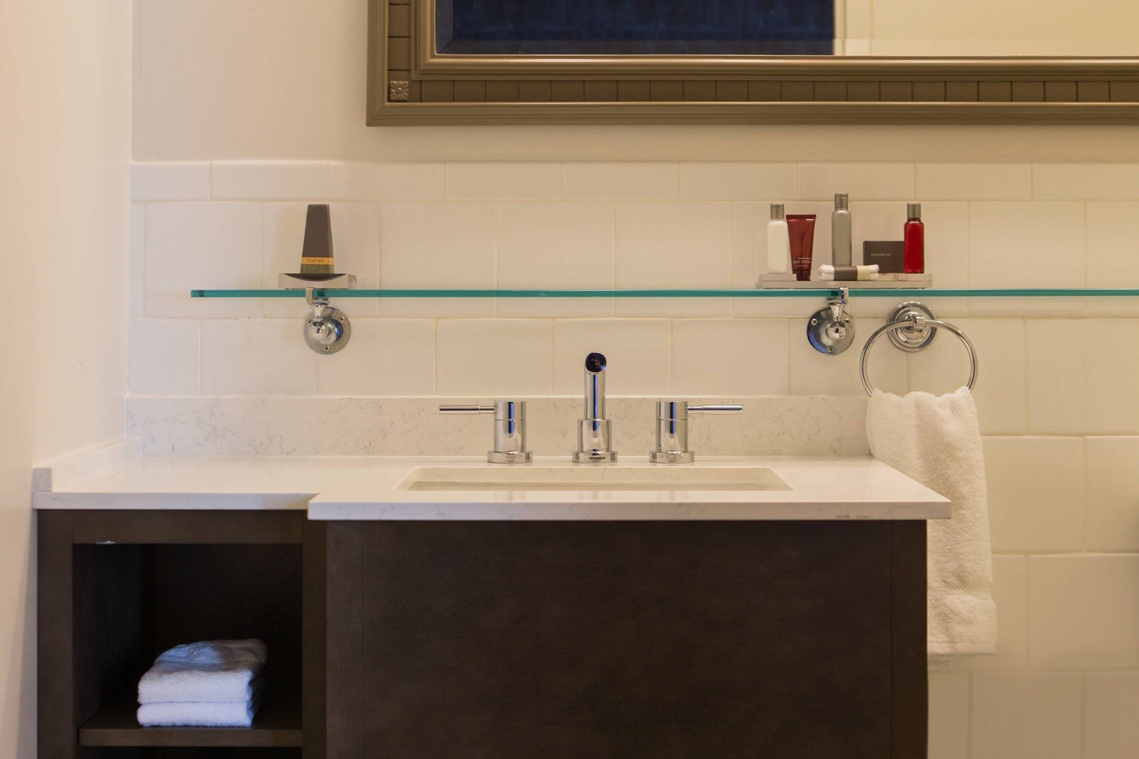 Une salle de bain avec un lavabo blanc, deux robinets, une étagère en verre où sont rangés les articles de toilette et un porte-serviettes.