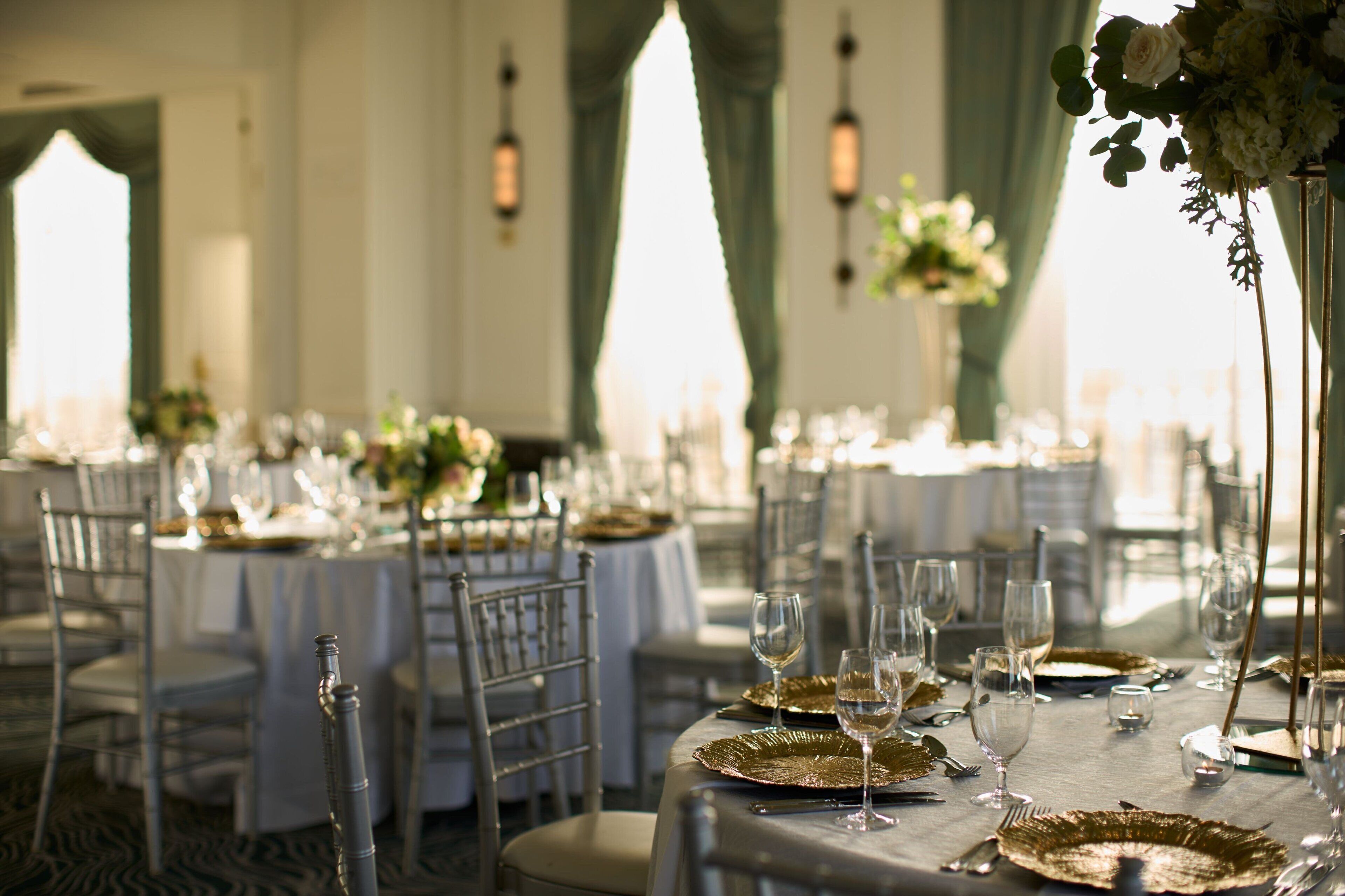 Uma sala de banquetes com mesas redondas dispostas para refeições, cada uma com um prato de apoio dourado, taça de vinho e talheres.