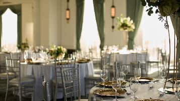 Uma sala de banquetes com mesas redondas dispostas para refeições, cada uma com um prato de apoio dourado, taça de vinho e talheres.