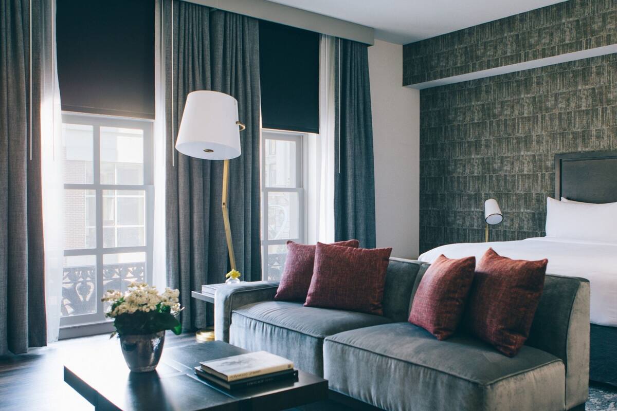 A modern hotel room with a grey sofa, a bedside table with a lamp, and a large window with curtains.