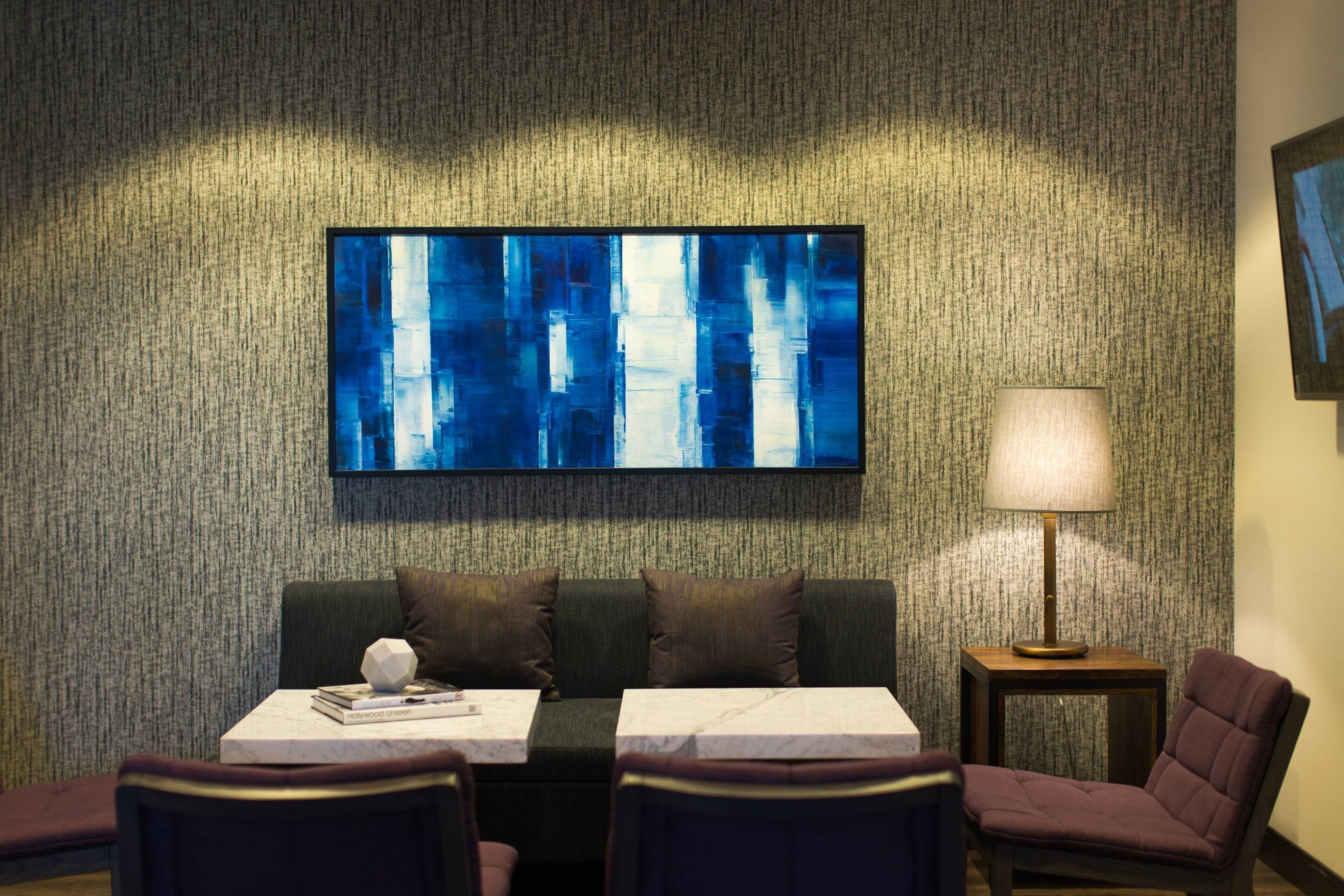 A modern hotel room with a textured wall, a large abstract painting, a sofa, two armchairs, a small table with a lamp, and a framed picture on the wall.