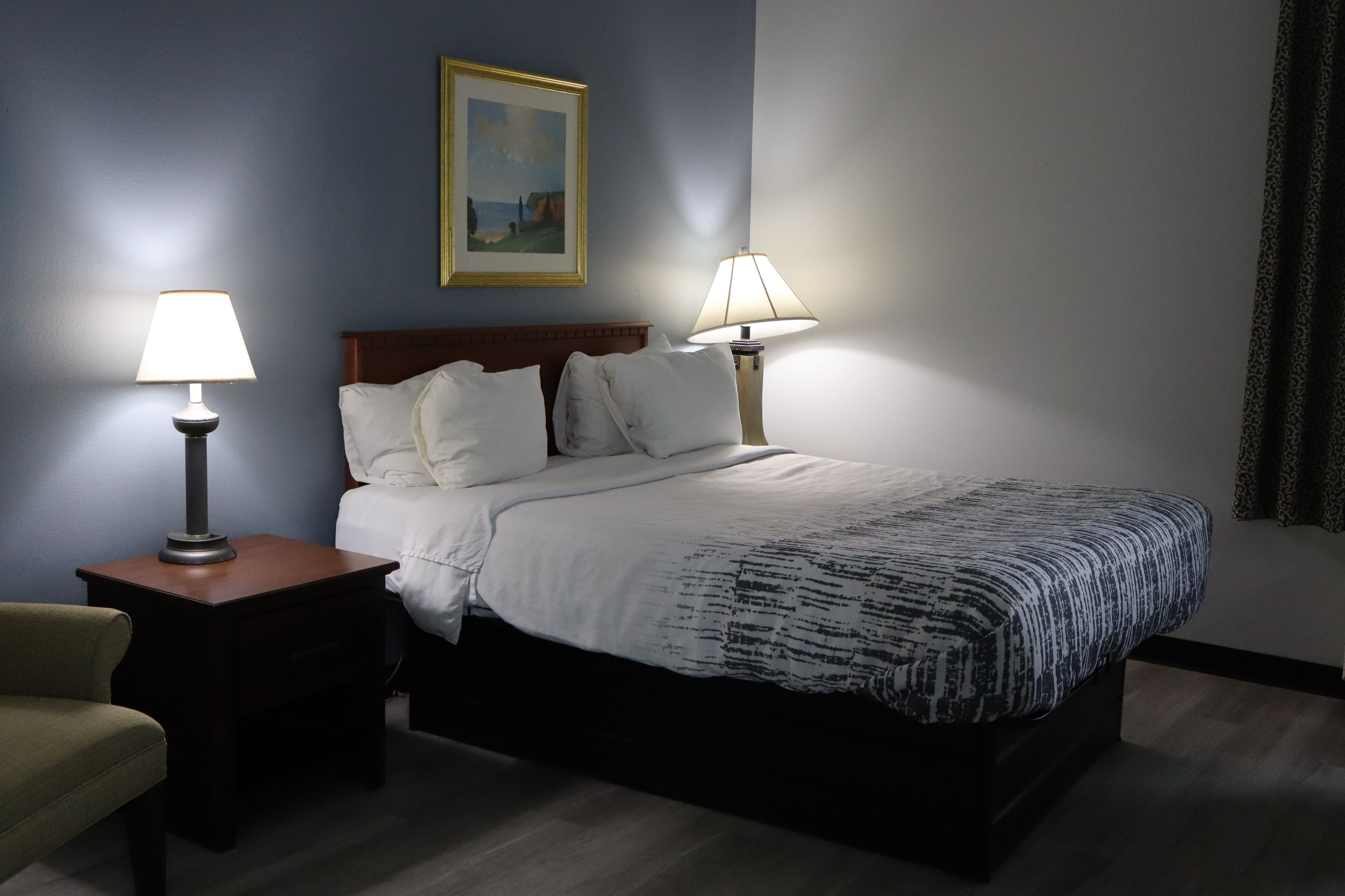 A hotel room with a bed, two bedside lamps, a chair, a framed picture on the wall, and a window with curtains.
