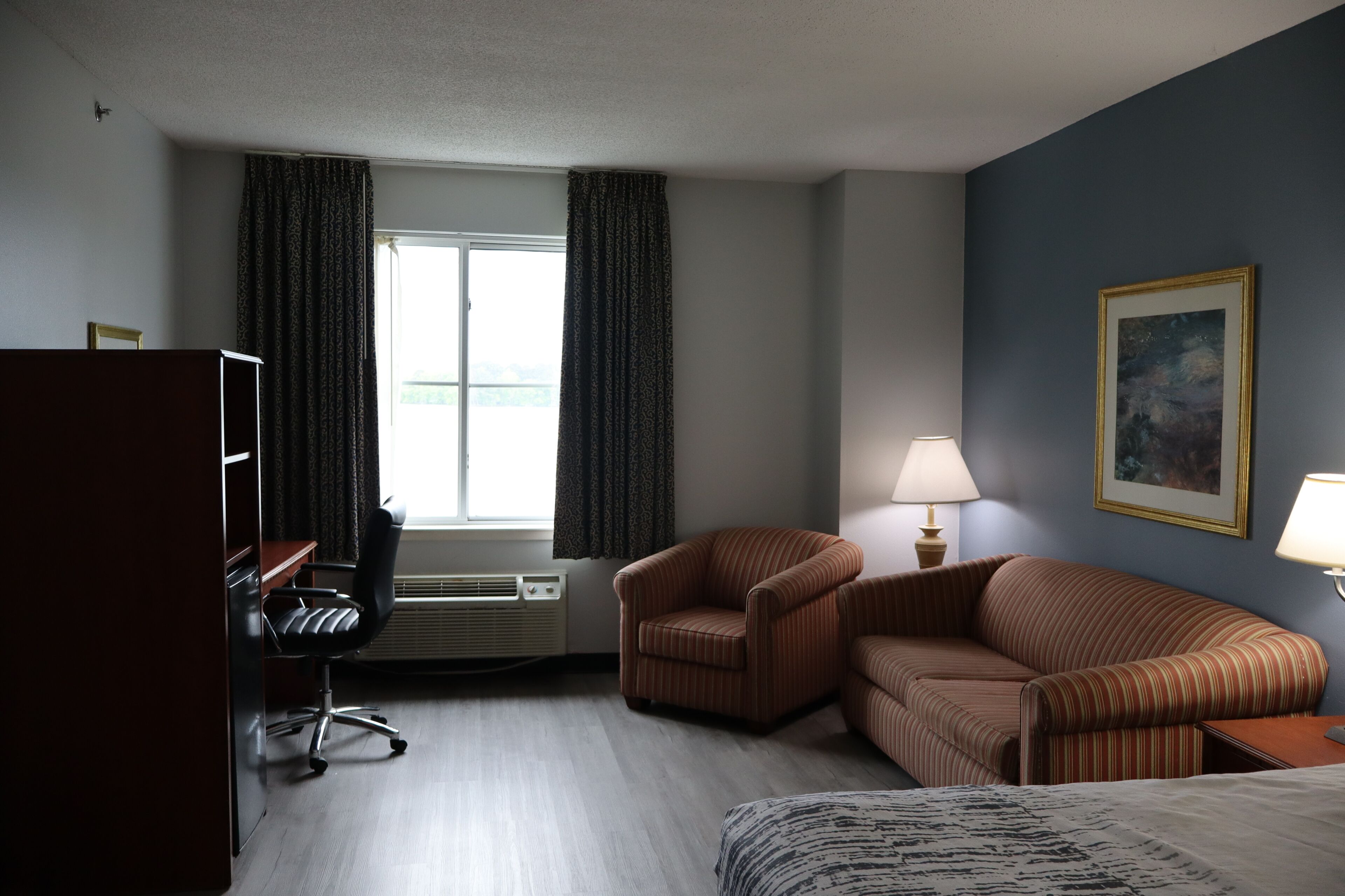 A hotel room with a desk, two chairs, a sofa, a bed, a window with curtains, a painting on the wall, and a lamp.