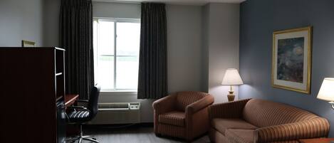 A hotel room with a desk, two chairs, a sofa, a bed, a window with curtains, a painting on the wall, and a lamp.