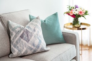 A grey sofa with patterned and solid teal cushions, a small round side table with a vase of flowers, and a white wall in the background.