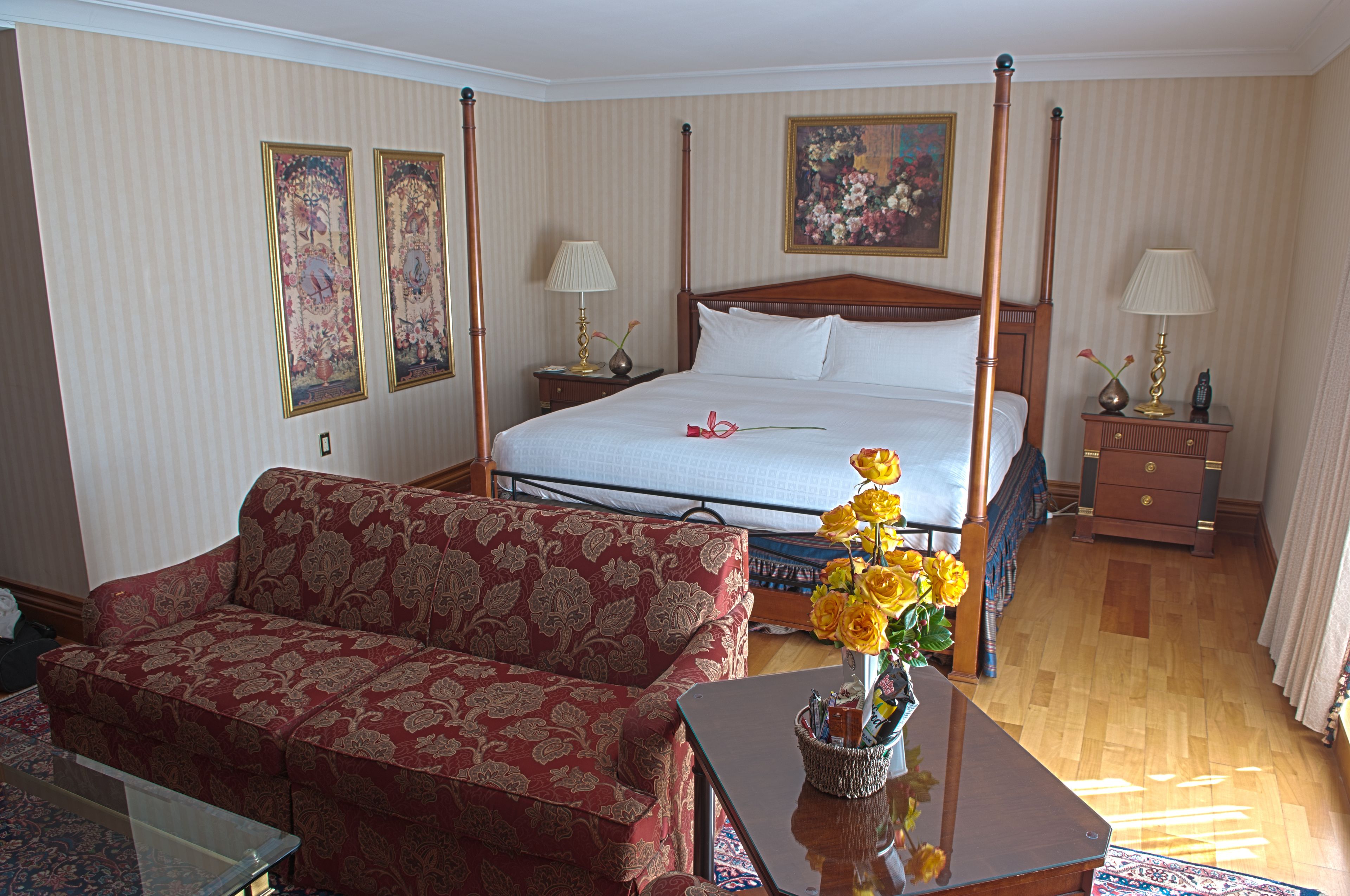 A hotel room with a four-poster bed, a red patterned sofa, a wooden coffee table with a vase of yellow flowers, and a small table with a lamp.