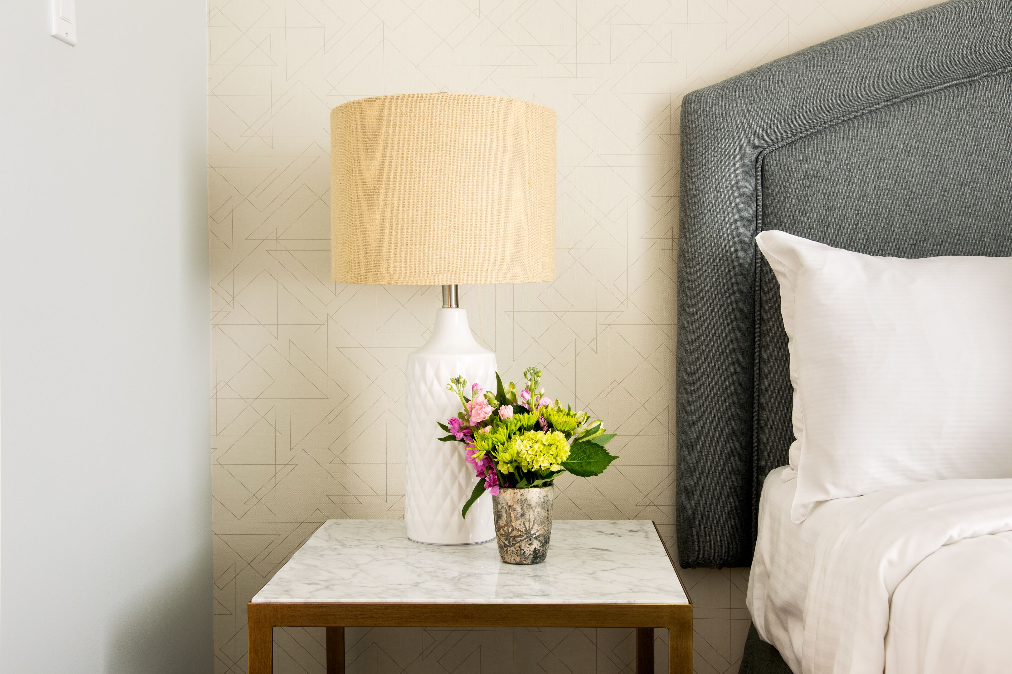 Habitación de hotel ordenada con cama, mesita de noche con lámpara y un jarrón con flores.