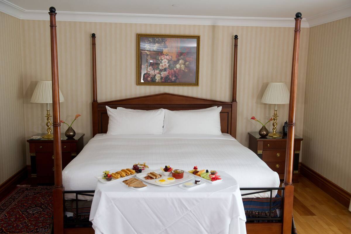 A four-poster bed with a white bedspread, a breakfast tray with eggs, toast, and fruit, and a painting above the bed.