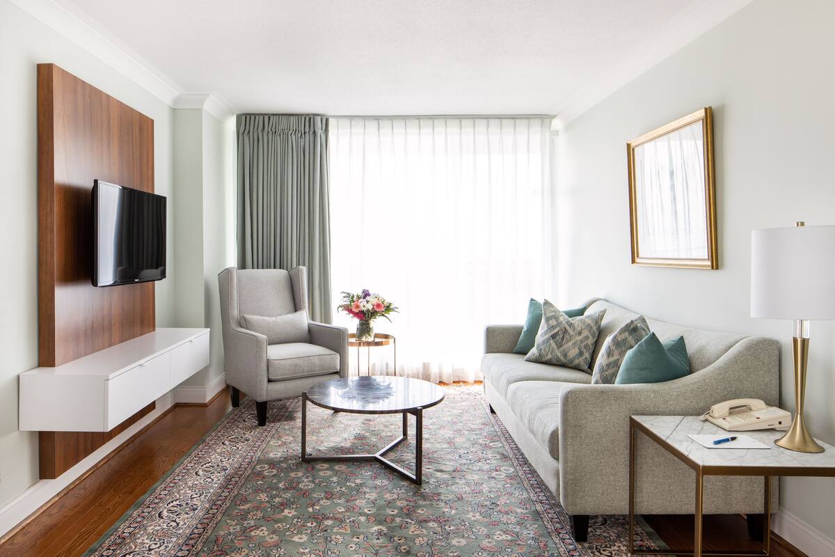 A modern living room with a flat-screen TV, a grey sofa with patterned cushions, a round coffee table, and a side table with a lamp and a telephone.