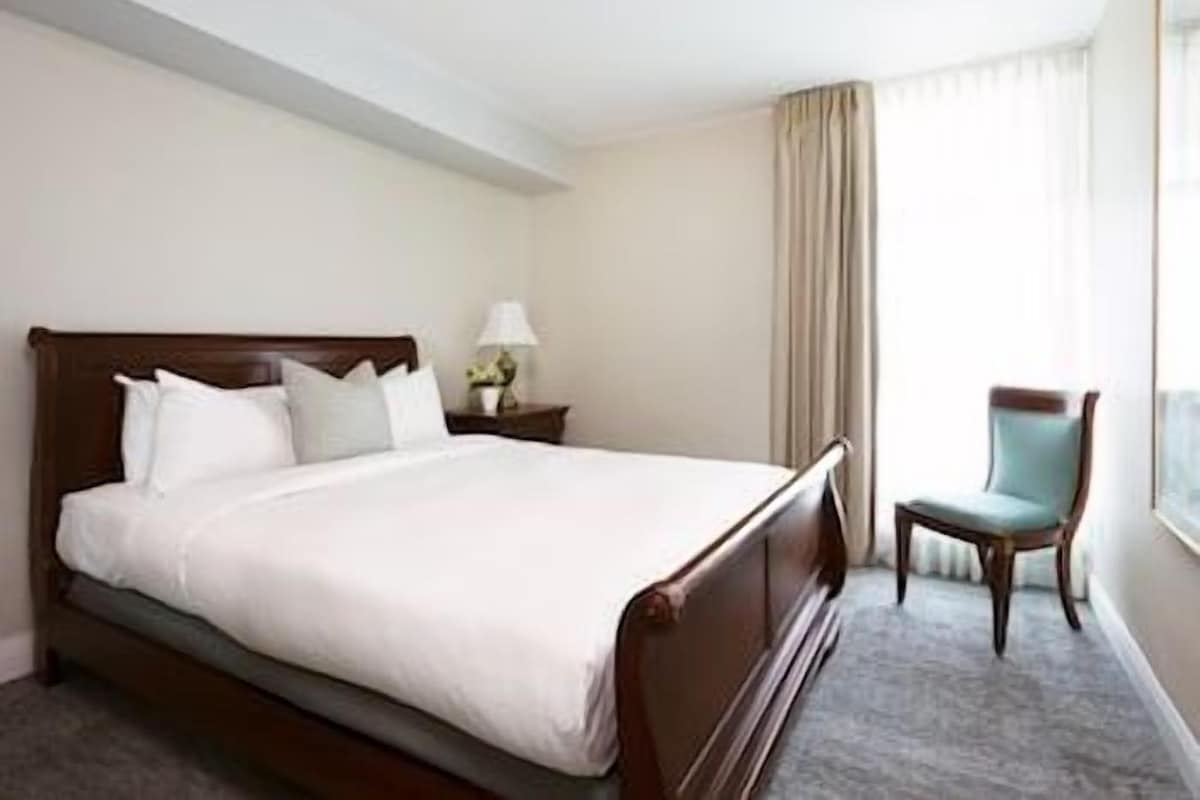 Quarto de hotel com uma cama grande, mesa de cabeceira com luminária, uma cadeira e uma janela com cortinas.