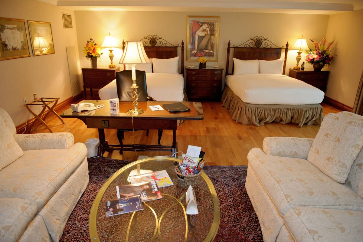 A hotel room with two beds, a desk with a lamp, a chair, a coffee table with magazines, and a vase with flowers.