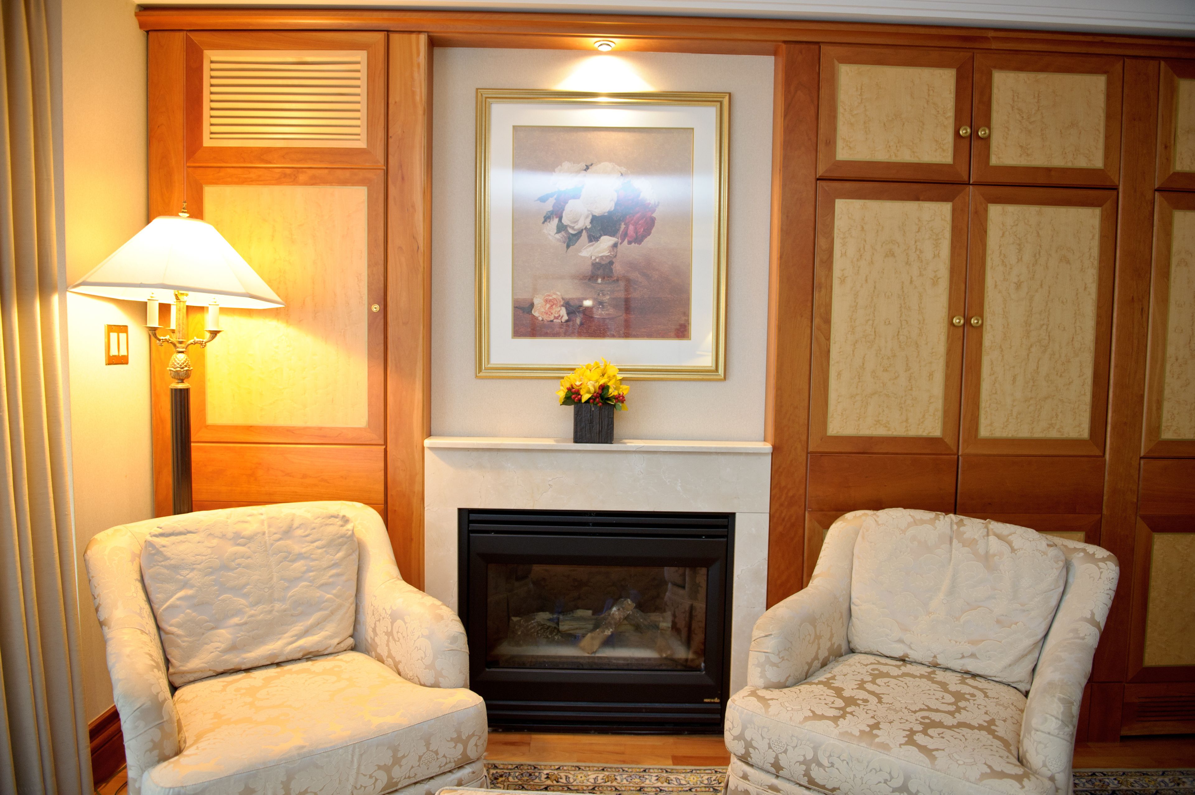 A room with a fireplace, two armchairs, a lamp, and a framed floral artwork above the fireplace.