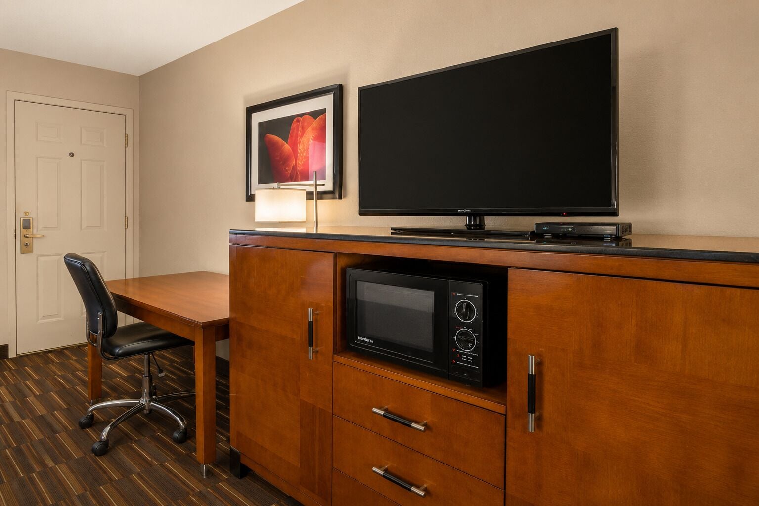A hotel room with a wooden desk, a black chair, a flat-screen TV, a microwave, and a framed picture on the wall.