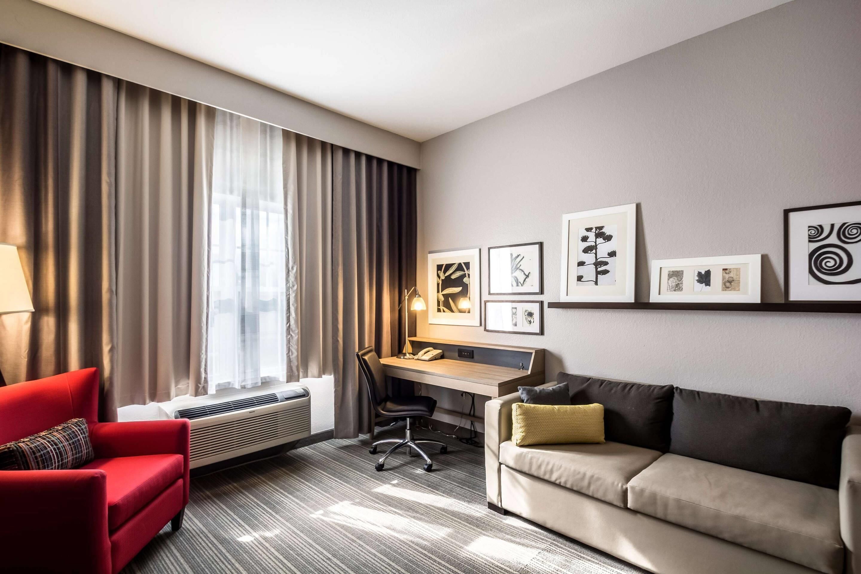 A hotel room with a red armchair, a beige sofa, a desk with a chair, and a large window with curtains.