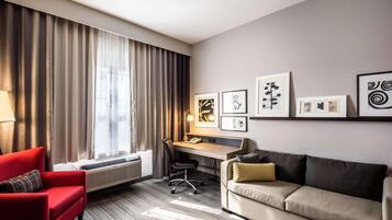 A hotel room with a red armchair, a beige sofa, a desk with a chair, and a large window with curtains.