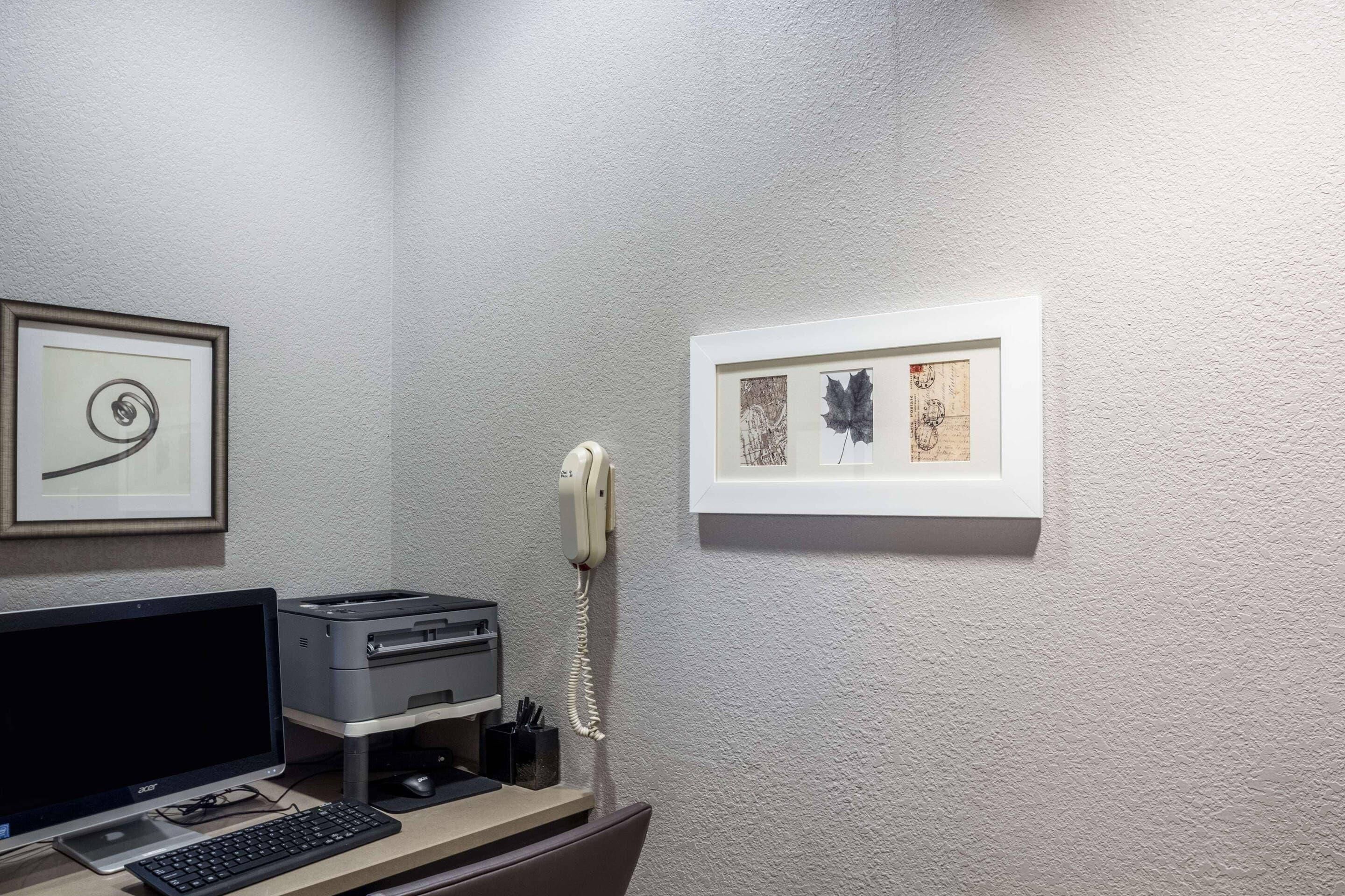 Ein Zimmer mit Schreibtisch, Desktop-Computer, Drucker, Telefon und gerahmten Bildern an der Wand.