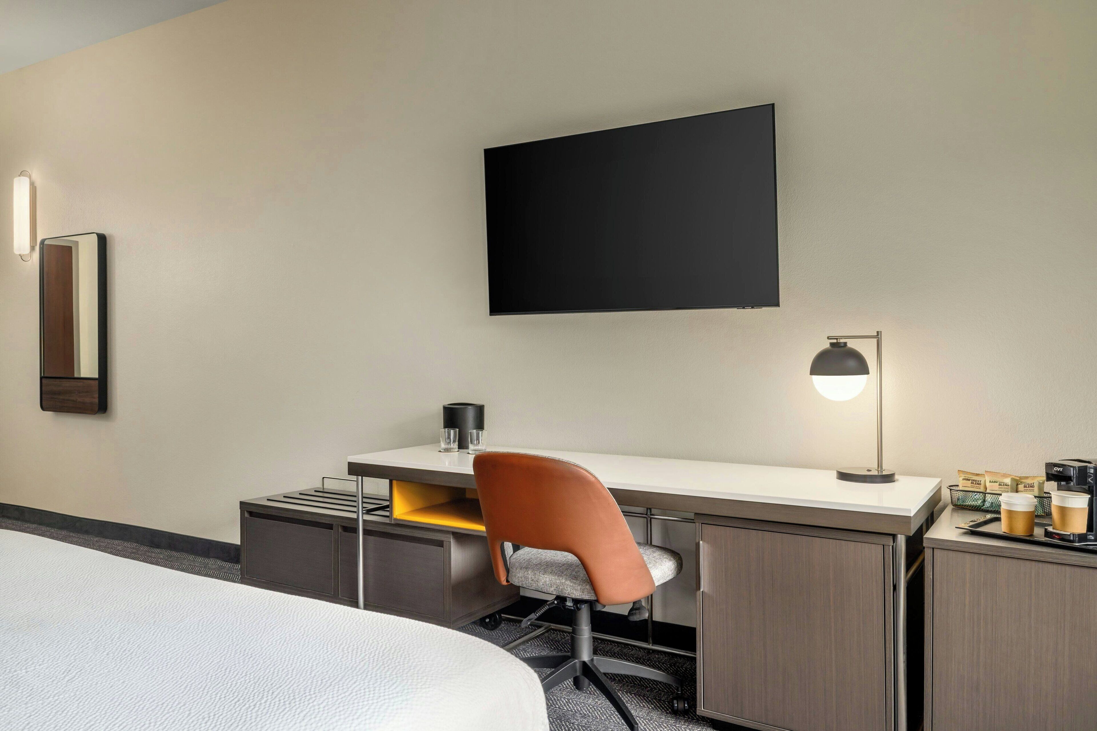 Habitación de hotel con cama, escritorio con silla, televisor de pantalla plana, lámpara y cafetera.
