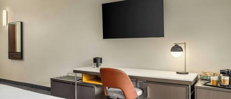 Habitación de hotel con cama, escritorio con silla, televisor de pantalla plana, lámpara y cafetera.