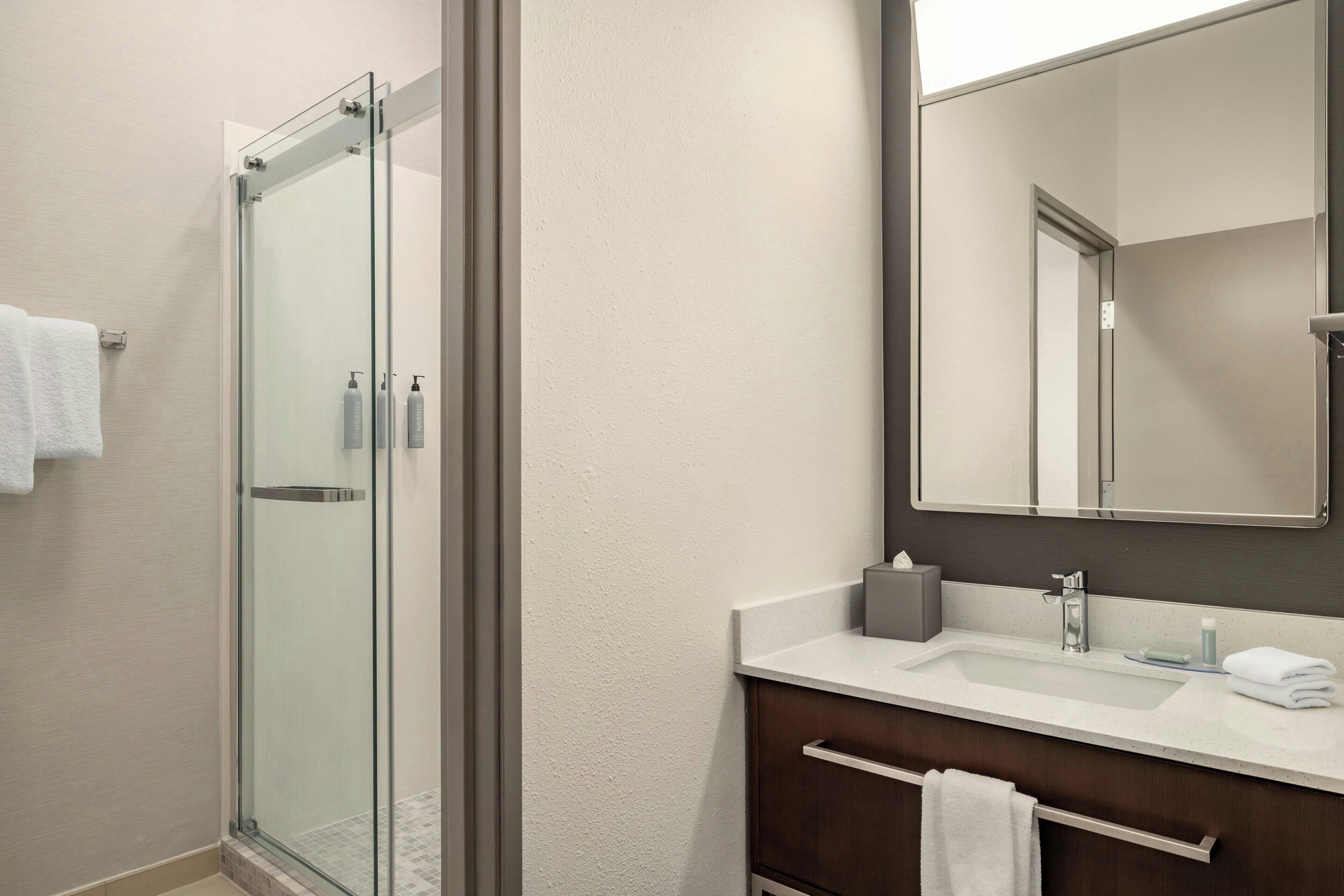Un baño moderno con ducha envuelta en vidrio, un espejo grande y un lavamanos blanco sobre un mueble de tocador oscuro.