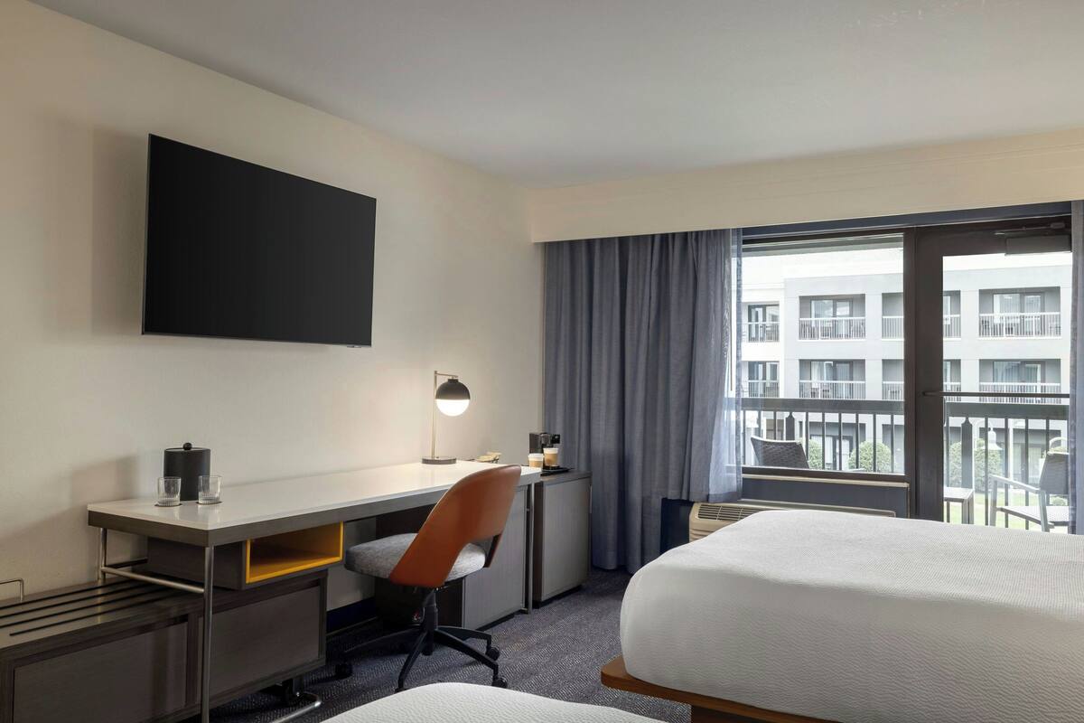 A hotel room with a bed, a desk with a chair, a flat-screen TV, a coffee machine, and a window with a view of a building.