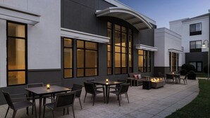 En udendørs terrasse med borde og stole, en bålende og en bygning med store vinduer.