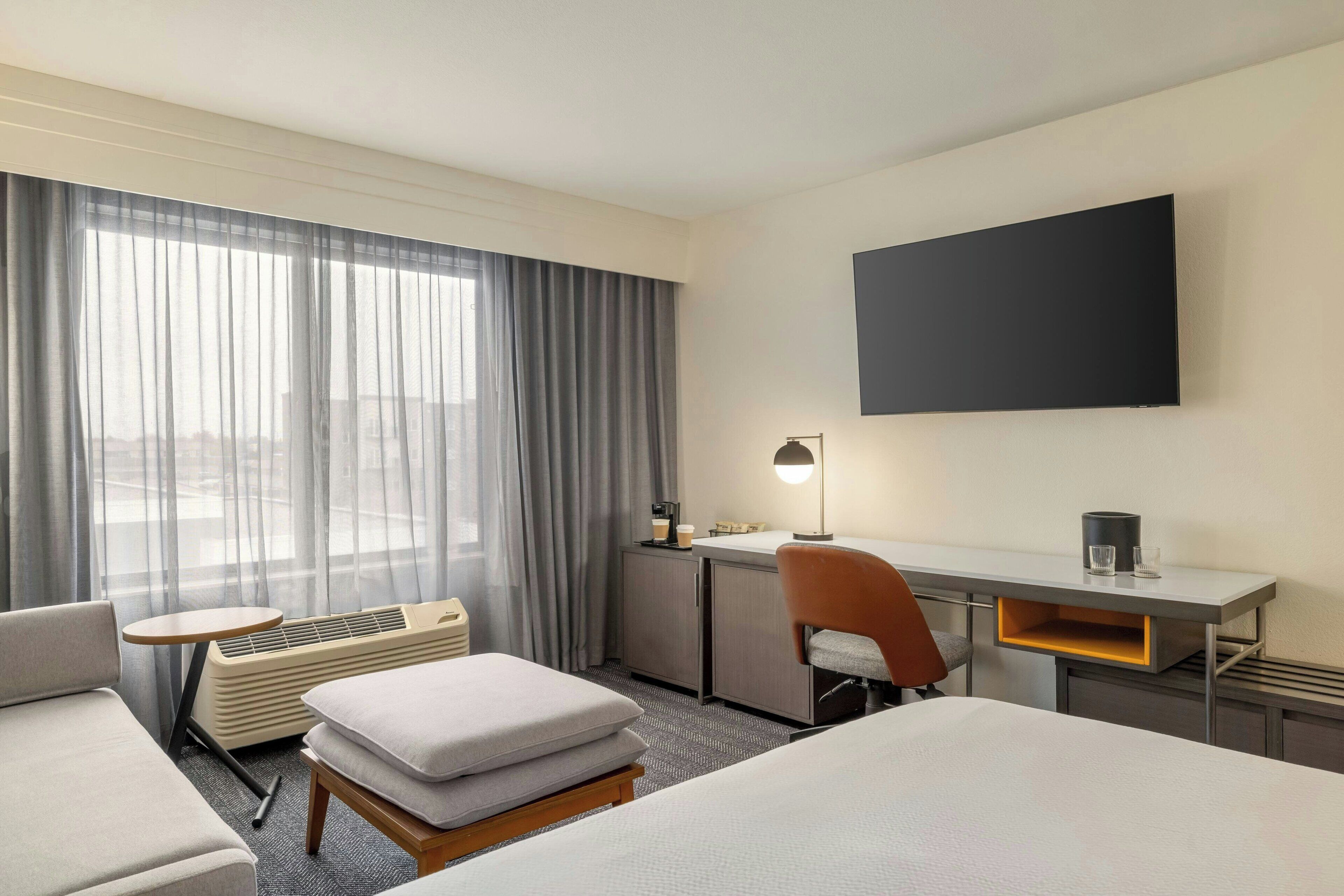 A hotel room with a bed, a desk, a chair, a small table, and a large window with curtains.